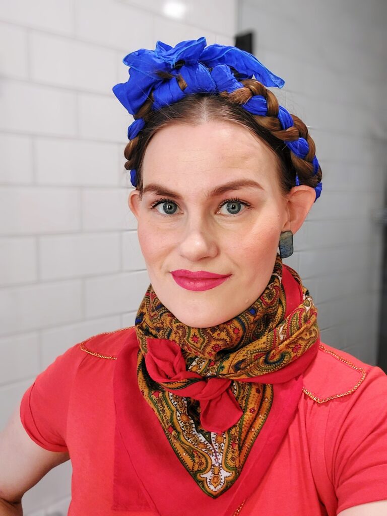 A woman with blue braided hair adorned with a matching bow, wearing a vibrant red top, ornate scarf, and statement earrings, smiles at the camera in a bright setting.