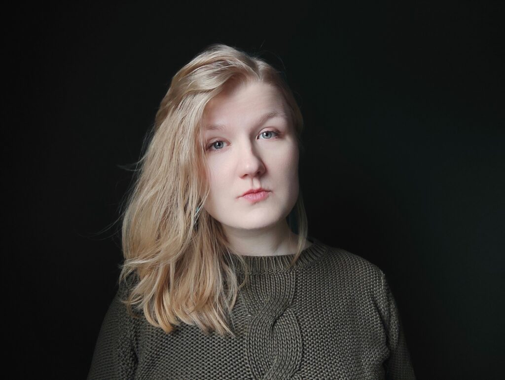 A contemplative young woman with blonde hair, set against a dark background, her gaze cast off to the side with a serene expression.