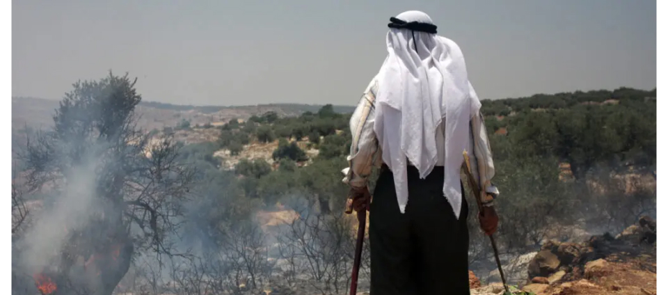 A person wearing traditional attire stands in a barren landscape, holding sticks in both hands. Smoke rises from small fires around them, and hills with scattered trees are visible in the background under a hazy sky.