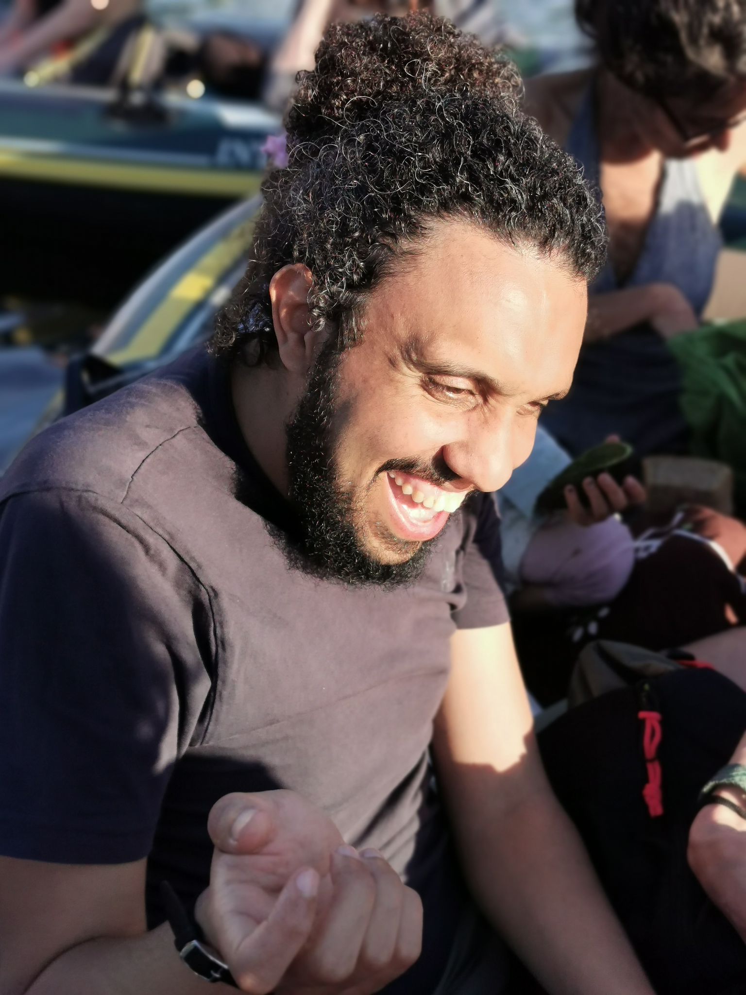 A man with curly hair and a beard, wearing a dark t-shirt, laughs joyfully while seated outdoors. Sunlight brightens his face, highlighting his smile. People and objects are blurred in the background.