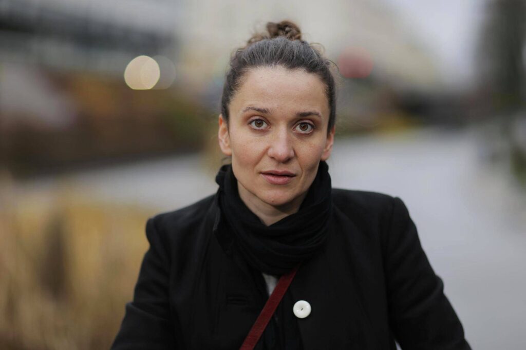 A person with long hair tied up, wearing a dark coat and scarf, stands outdoors with a blurred cityscape background. Their expression is neutral, and they are facing the camera directly.