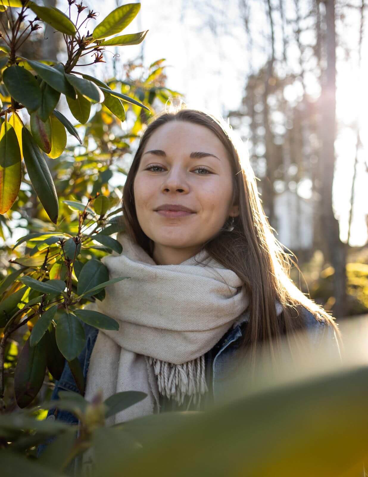 A young woman with long brown hair wearing a beige scarf stands among green foliage on a sunny day. Sunlight filters through the trees in the background, creating a warm and serene ambiance. She is slightly smiling and looking directly at the camera.