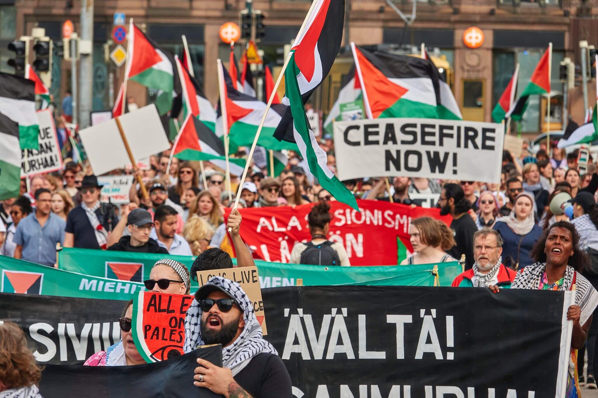 A large crowd of protestors is marching with various signs and banners, including messages like "Ceasefire Now!" and displaying Palestinian flags. The protest appears to be in a city setting with buildings and transport visible in the background.